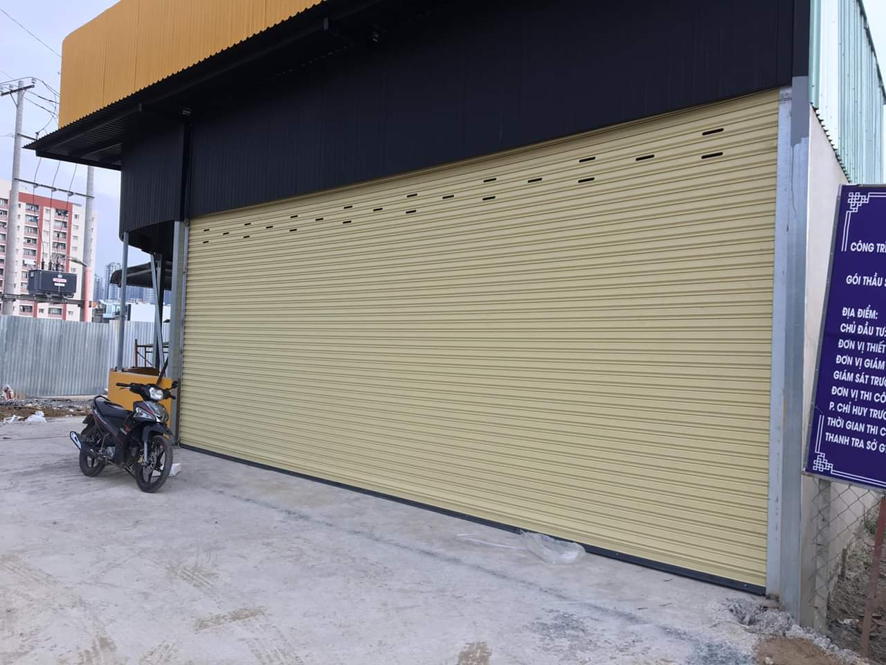 Cửa cuốn tôn màu vàng lắp cho mặt tiền nhà phố, phía trước có xe máy dựng trên nền bê tông
