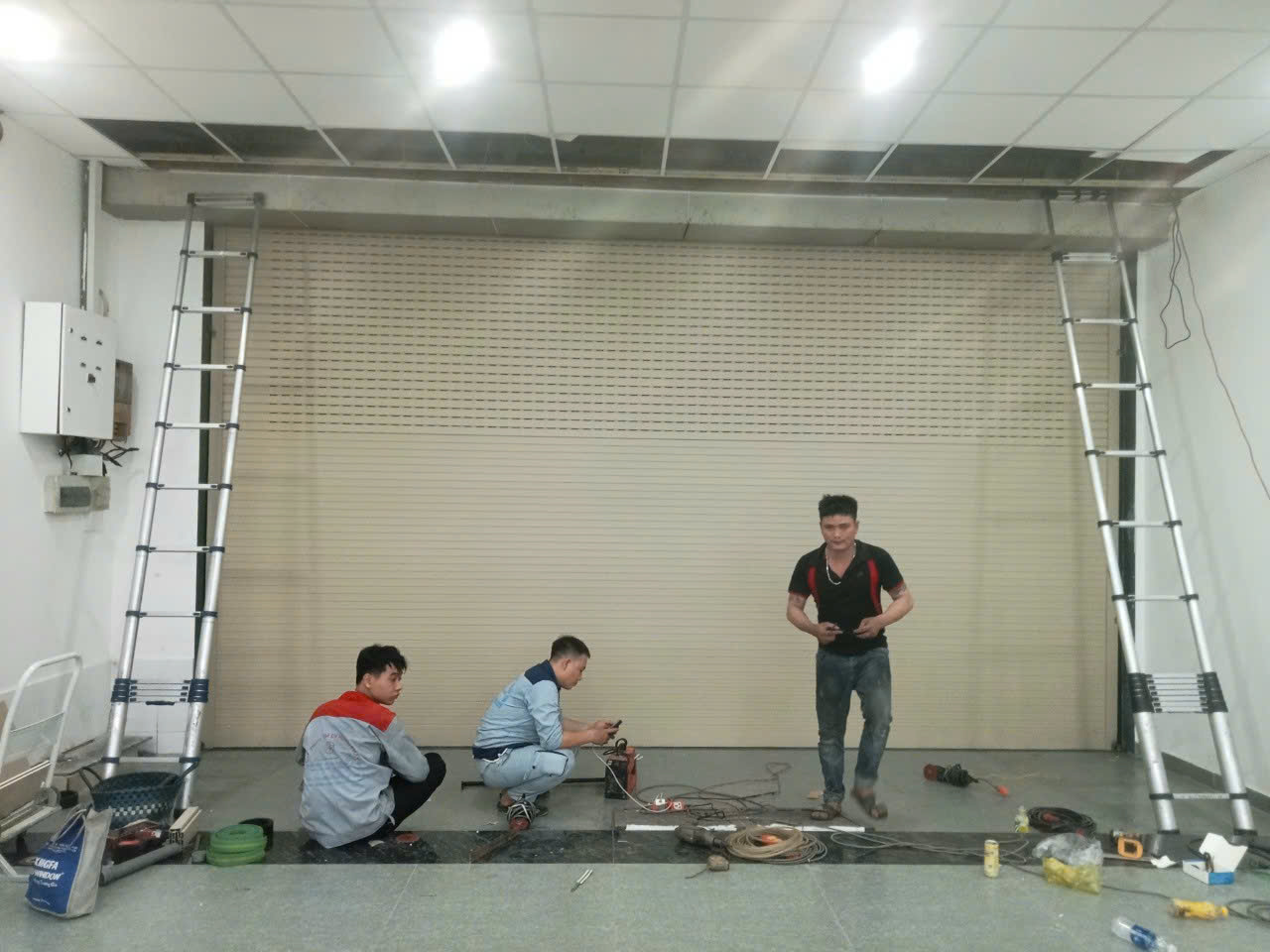 Thợ đang lắp đặt cửa cuốn tự động màu kem trong nhà, xung quanh là thang nhôm và dụng cụ điện