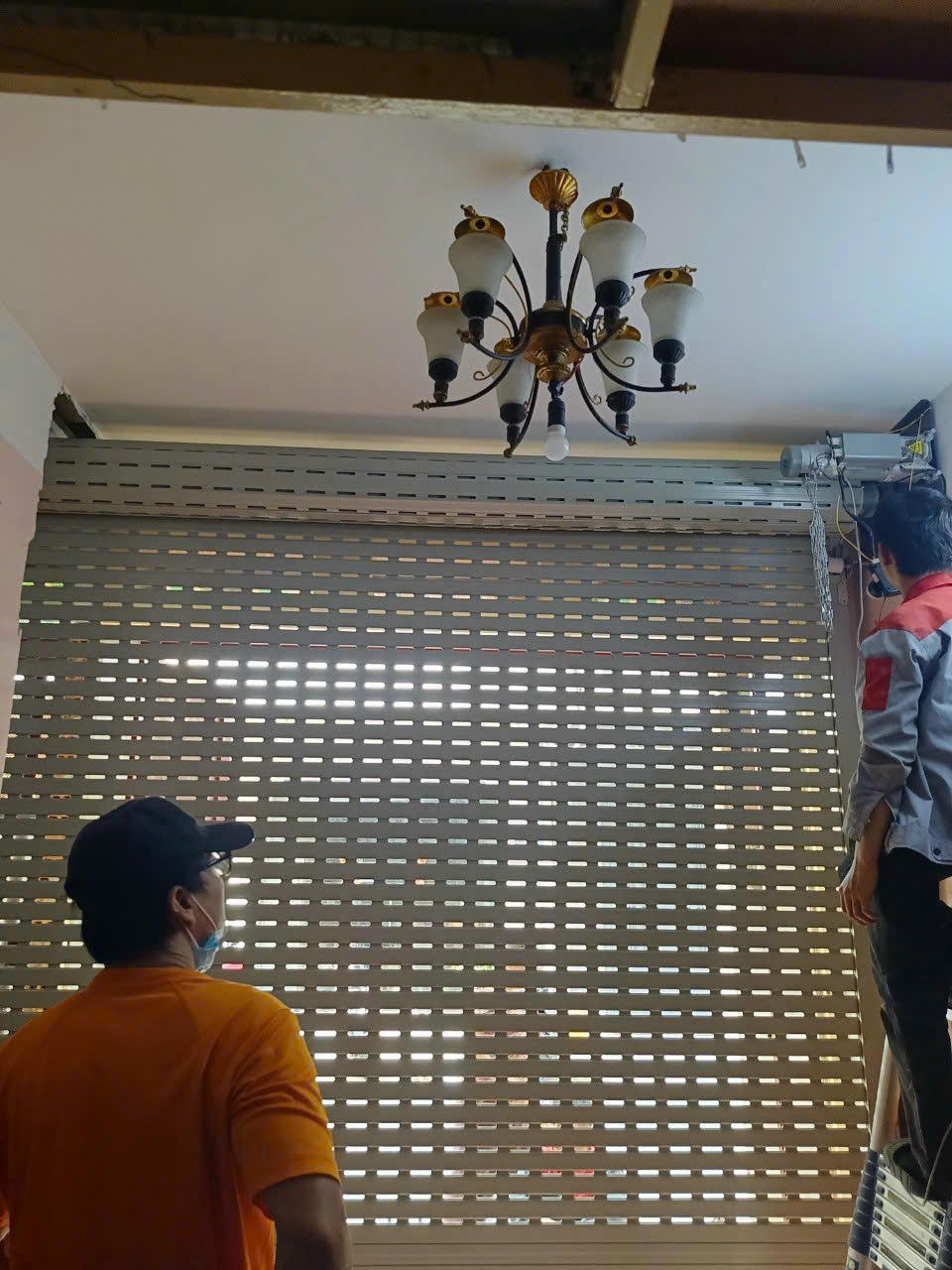 Thợ đang lắp đặt cửa cuốn khe thoáng trong nhà dưới đèn chùm trần