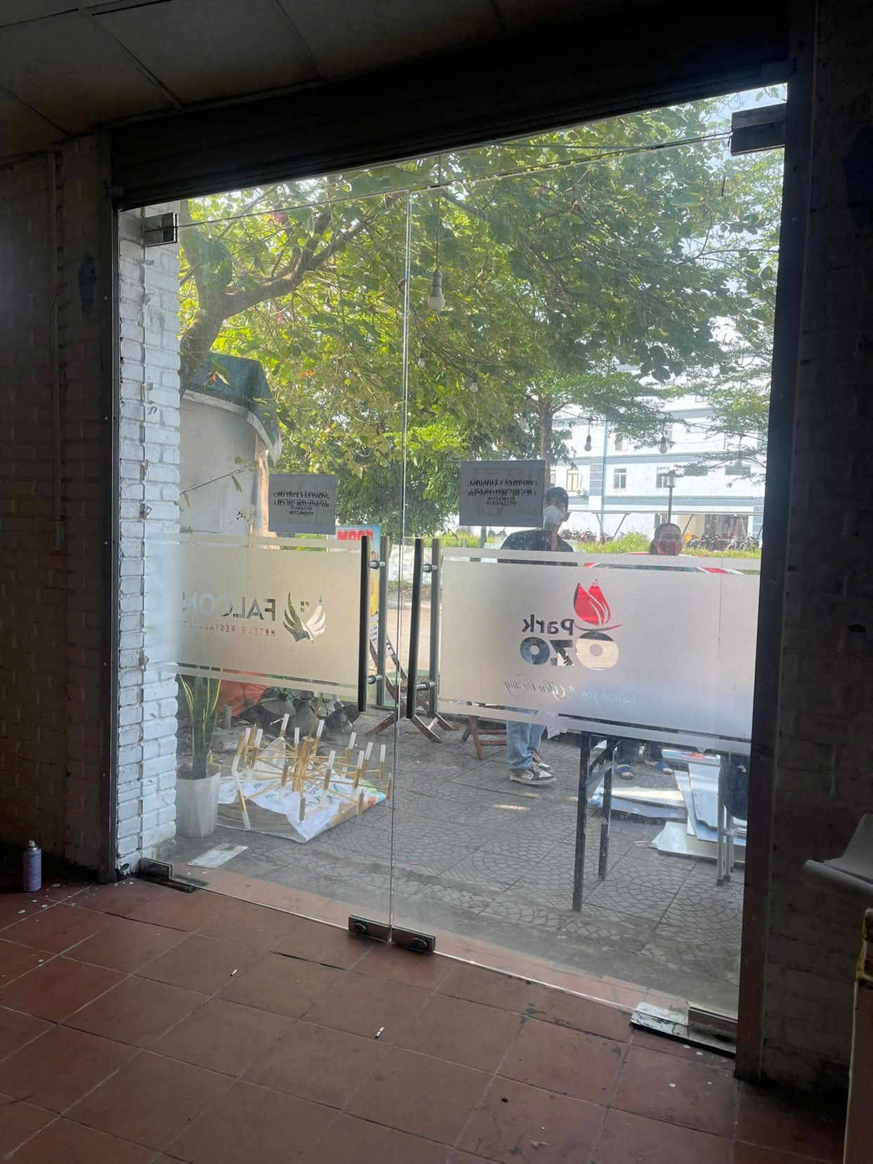 Cửa kính cường lực hai cánh có dán decal mờ logo quán nhìn ra vỉa hè và cây xanh