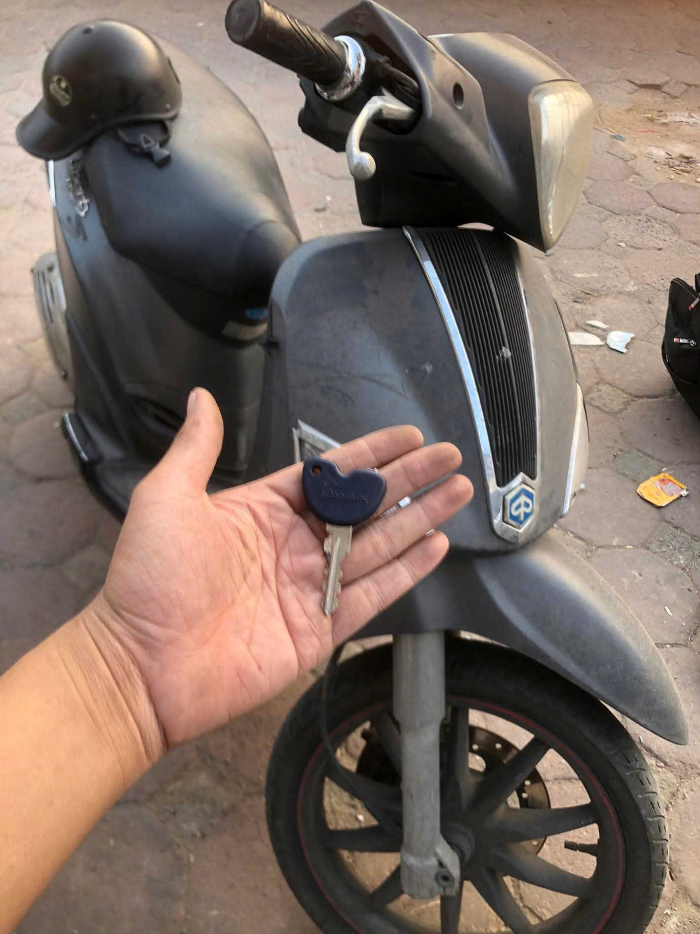 Chìa khóa xe máy vespa Piaggio trên tay người, phía sau là chiếc xe tay ga Piaggio màu xám