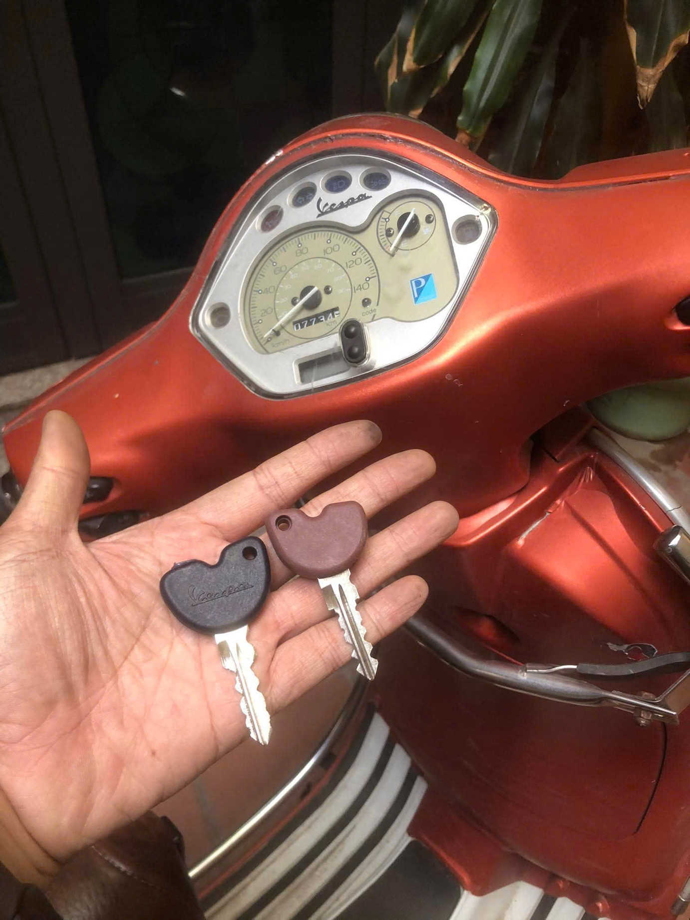 Chìa khóa xe Vespa trên tay trước bảng đồng hồ và thân xe máy Vespa màu cam
