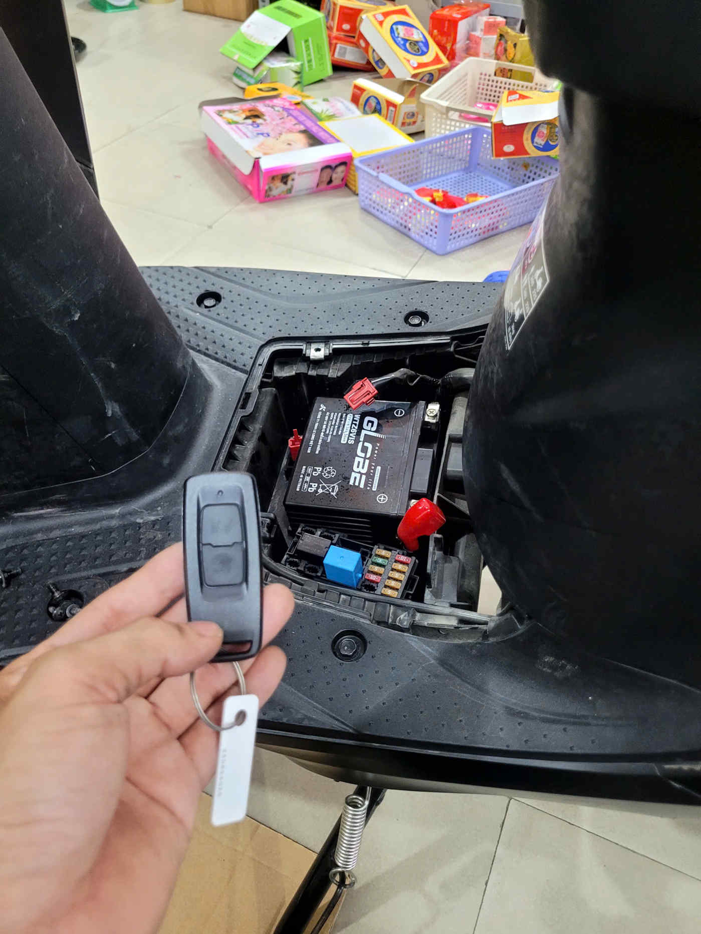 Chìa khóa remote xe máy điện cầm trên tay cạnh bình ắc quy và hộp cầu chì dưới sàn để chân