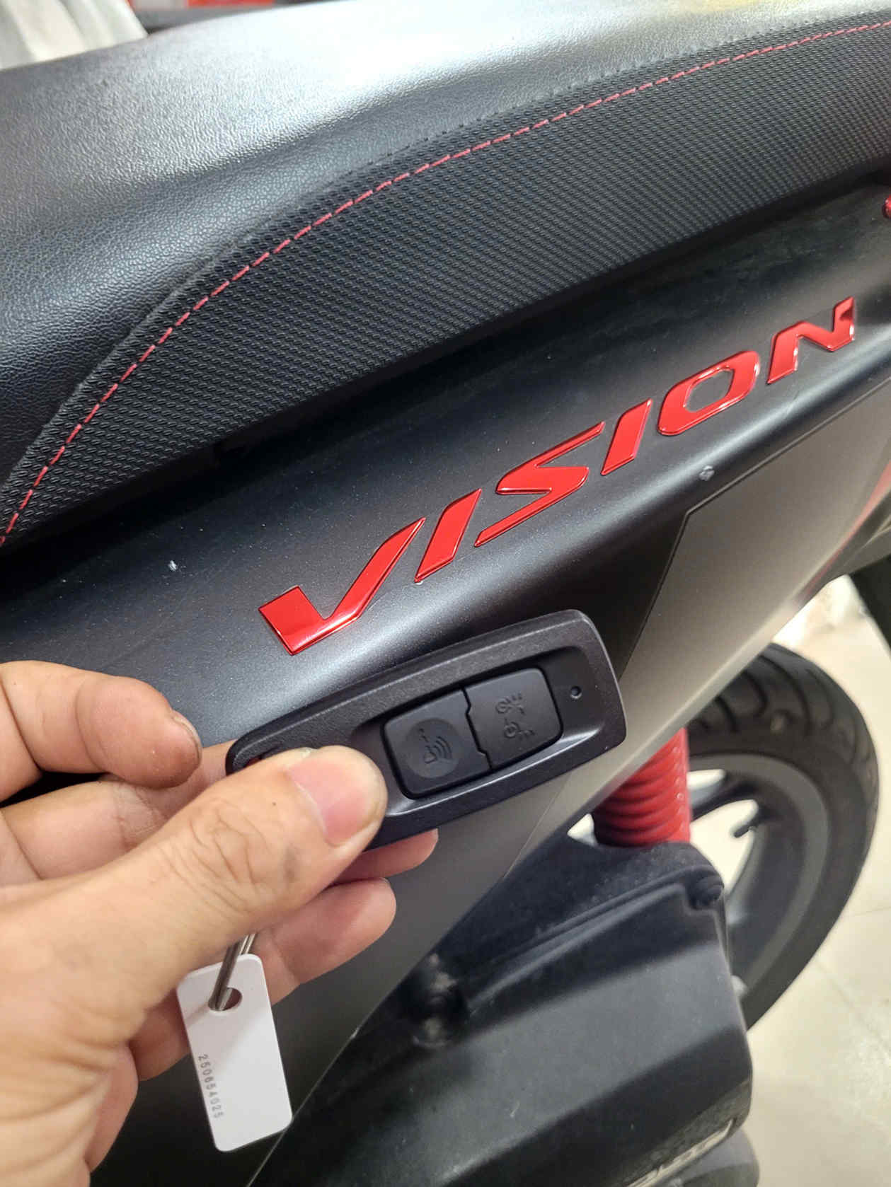 Chìa khóa thông minh xe máy Honda Vision màu đen cầm trên tay cạnh yên xe và logo Vision đỏ