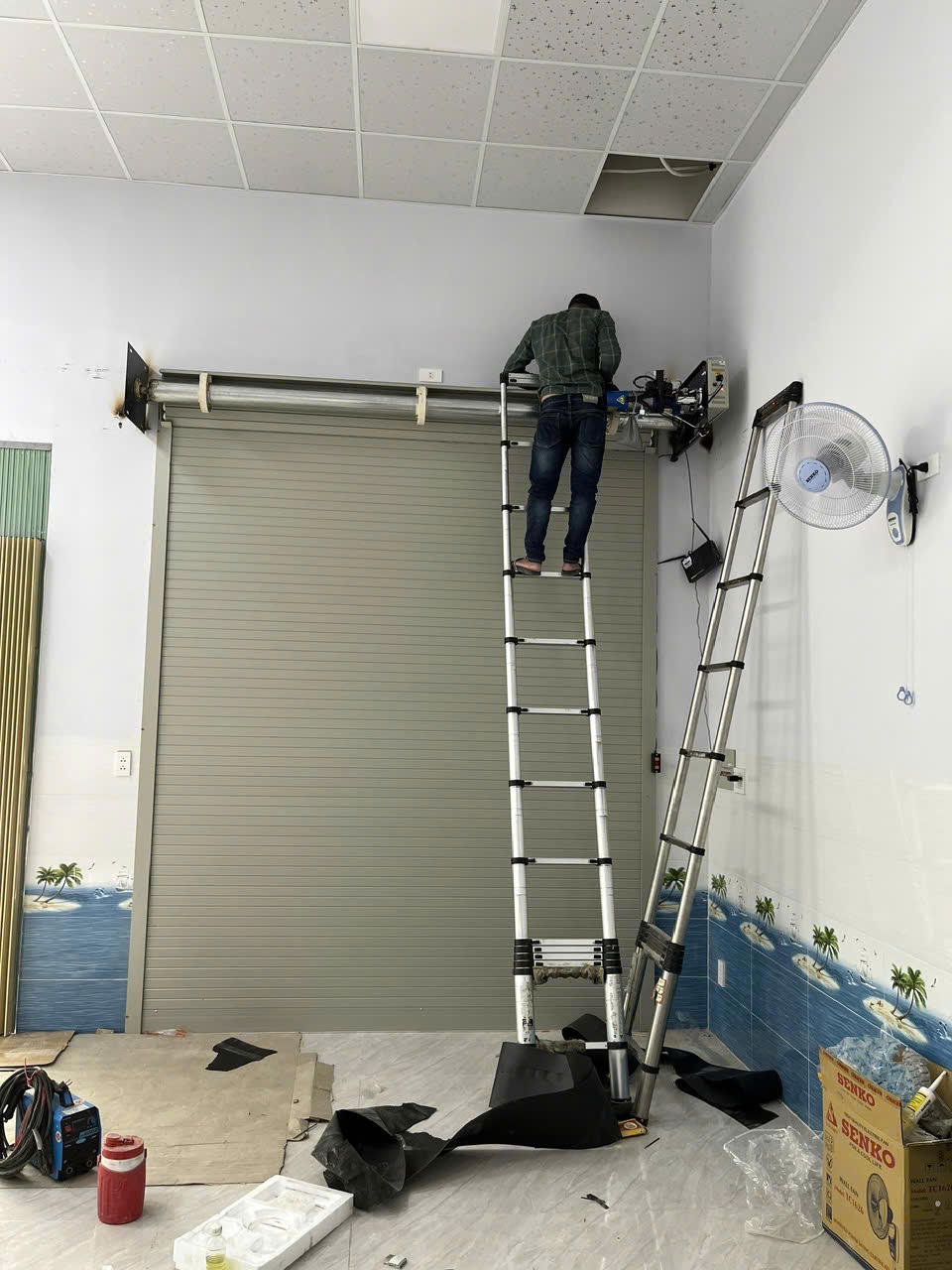 Thợ đang lắp đặt motor cửa cuốn tôn màu xám trong nhà với hai thang nhôm dựng sát tường