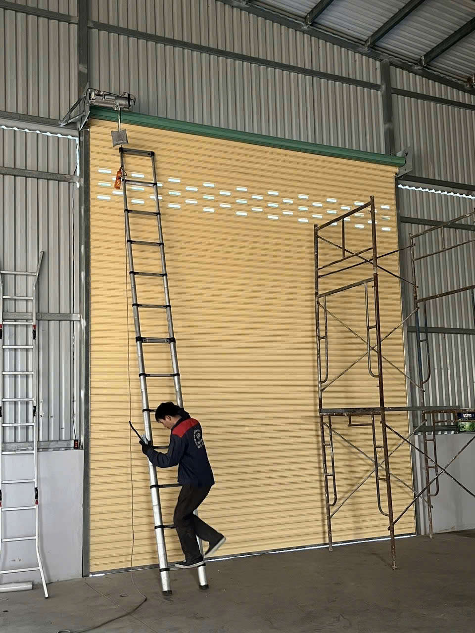 Thợ kỹ thuật lắp đặt cửa cuốn tôn màu vàng cho nhà xưởng khung thép