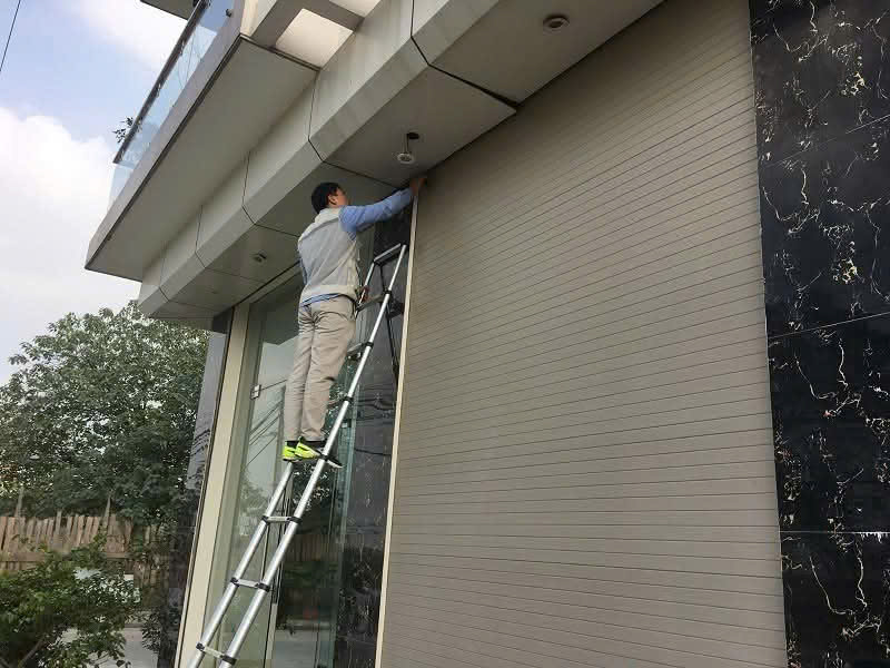 Kỹ thuật viên đứng trên thang lắp đặt cửa cuốn tự động cho mặt tiền nhà kính