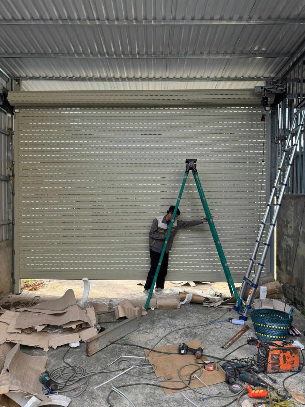 Thợ đang lắp đặt cửa cuốn tôn màu kem trong gara, xung quanh có thang và dụng cụ thi công