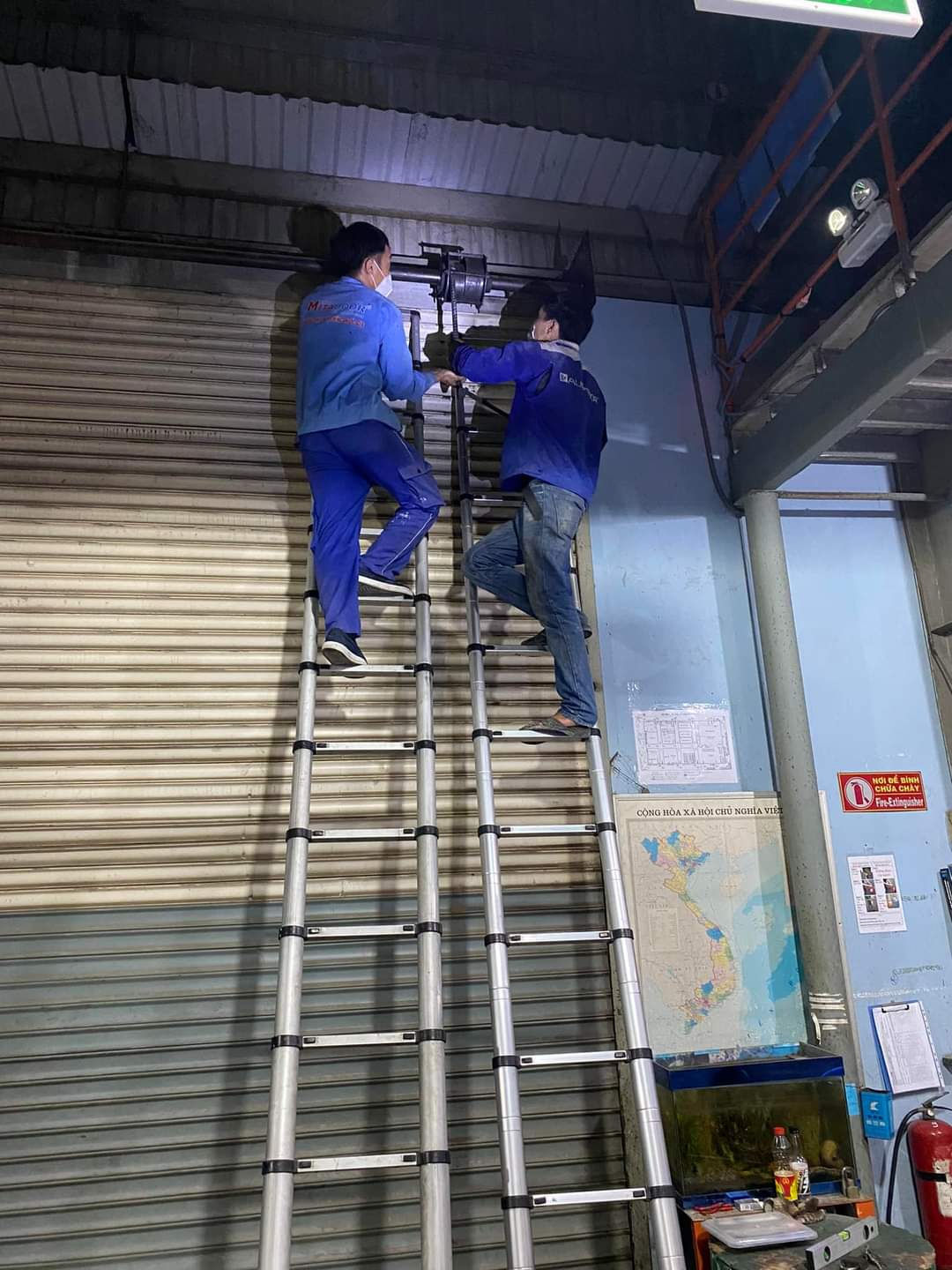 Thợ kỹ thuật đứng trên thang nhôm rút đôi sửa chữa motor cửa cuốn nhà xưởng