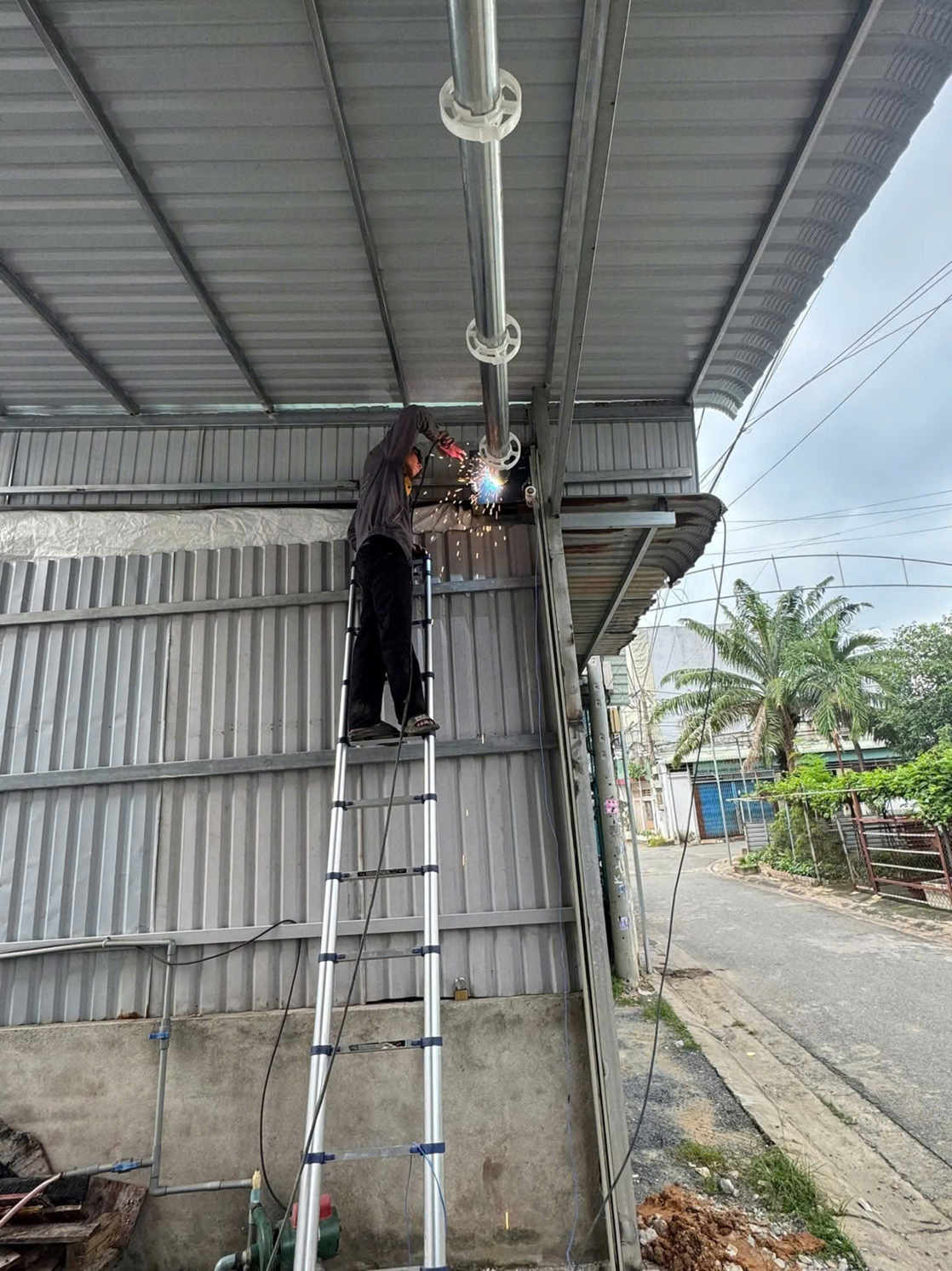 Thợ hàn đứng trên thang nhôm đang hàn sửa cửa cuốn bị gãy trục dưới mái tôn nhà xưởng ngoài trời