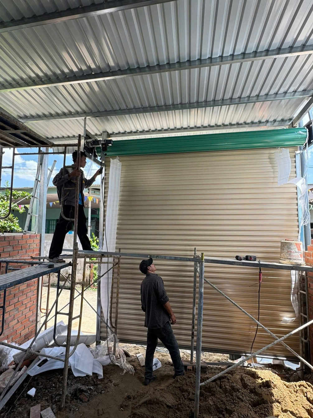 Thợ đang lắp đặt sửa cửa cuốn tôn màu kem cho mặt tiền nhà đang xây dựng