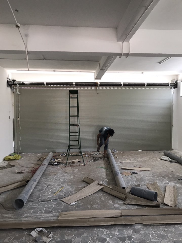 Công nhân lắp đặt cửa cuốn kim loại cho mặt bằng nhà phố, xung quanh còn nhiều vật liệu xây dựng