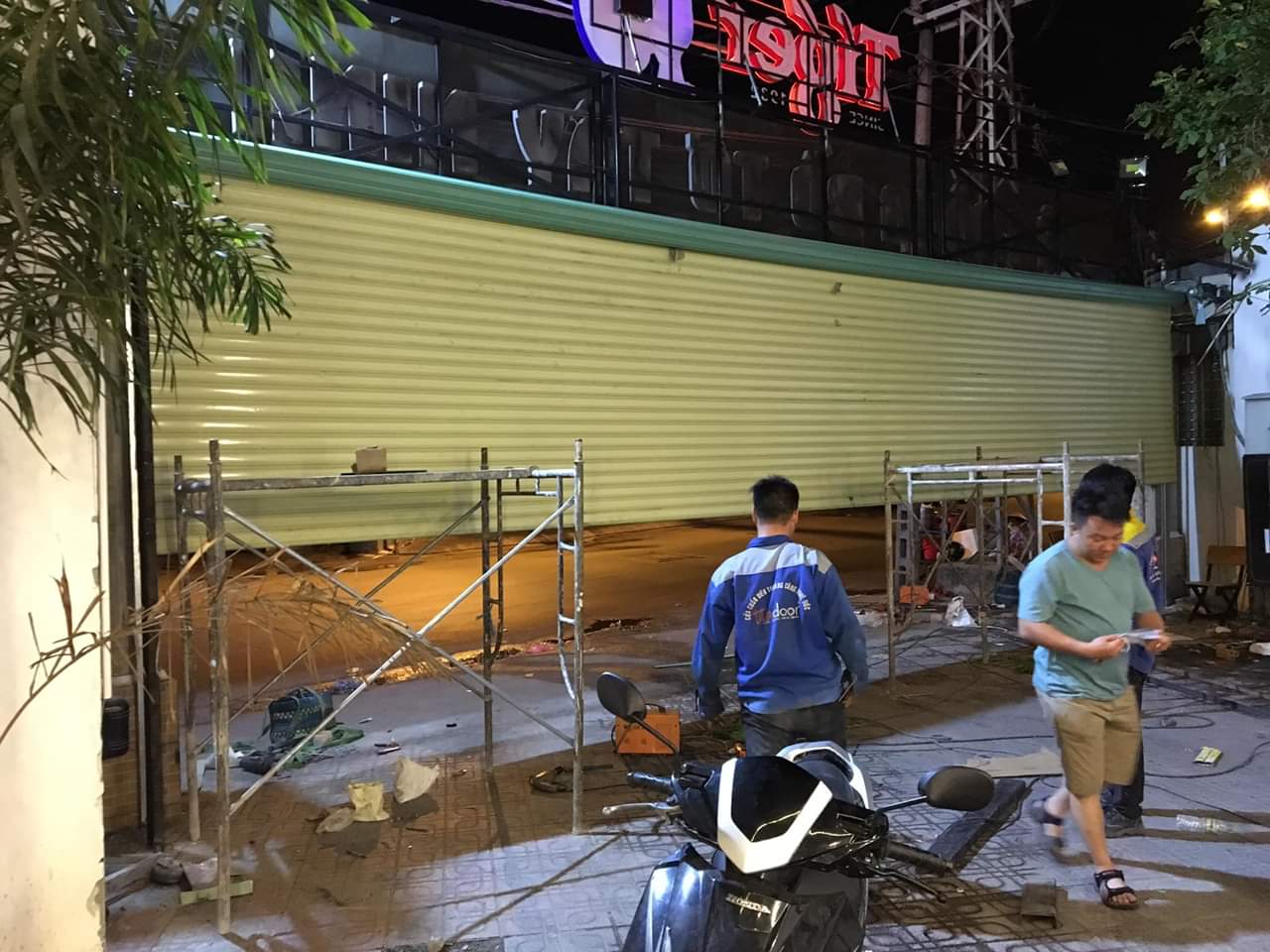 Cửa cuốn tôn màu vàng đang thi công buổi tối với giàn giáo và thợ lắp đặt trước mặt tiền cửa hàng